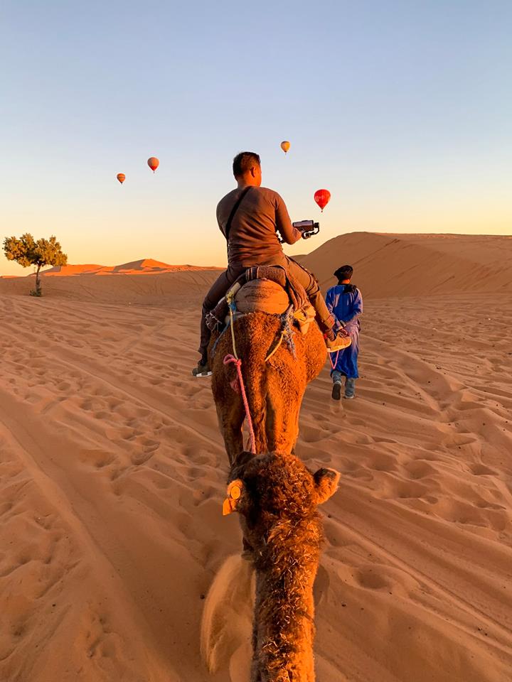 Riding the camel at Sahara Desert during sunset with hot air balloon as the backdrop